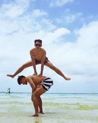 Full length of shirtless man jumping over friend bending on shore at beach against sky