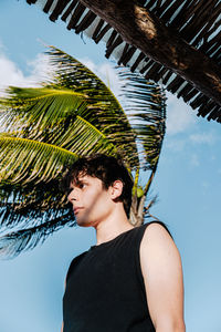 Portrait of young woman standing against palm tree against sky