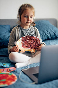 Full length of cute girl sitting on bed with laptop at home