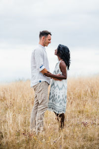 Side view of smiling man embracing indian girlfriend while looking at each other in field under cloudy sky