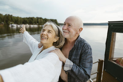 Senior man with hands on happy woman's shoulder on houseboat near river