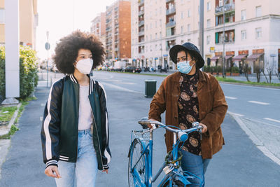 Female friends with protective face masks walking by bicycle on road