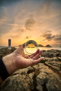 Person holding rock in sea against sky during sunset