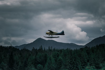 Low angle view of airplane flying over forest against cloudy sky