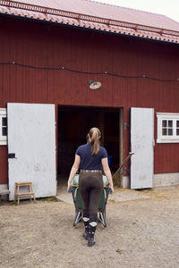 Rear view of female farmer pushing wheelbarrow towards barn