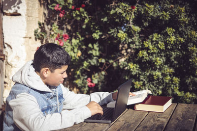 Young latin man reads book in front of laptop, outdoors