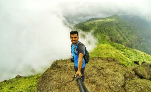 Smiling man standing on cliff taking selfie against cloudy mountains