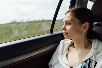 Pensive young female with dark hair in casual clothes sitting near window on backseat of modern car and looking during road trip on cloudy day
