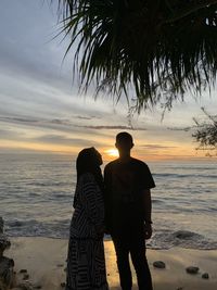 Rear view of friends standing at beach against sky during sunset