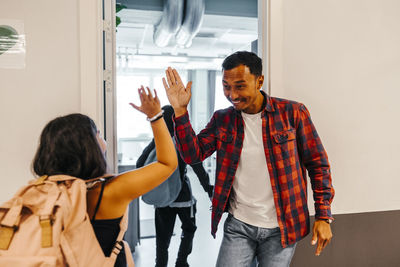 Cheerful male teacher giving high-five to female pupil while standing near doorway of classroom in school