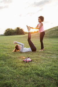 Girl helping mother exercising on grass at park against sky during sunset