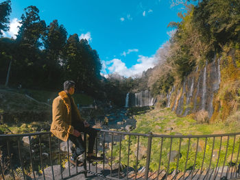 Man looking away while sitting on railing against trees