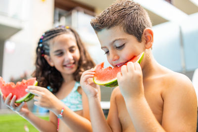 Two funny kids eating watermelon at the the edge of the pool on a summer day