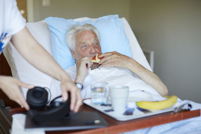 Senior man eating cracker sandwich while female nurse holding headphones in hospital ward