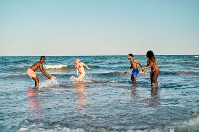Diverse young men and women in swimwear smiling and splashing water while having fun in waving sea on summer weekend day