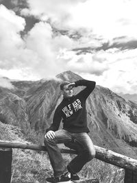 Man sitting on railing with mountains in background against cloudy sky