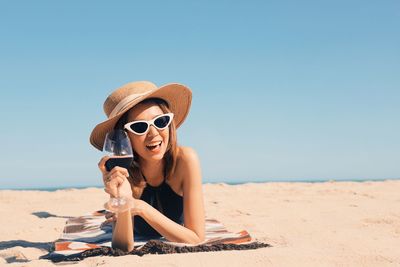 Young woman wearing sunglasses while sitting at beach against clear sky
