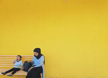 Mother and son sitting on bench against yellow wall
