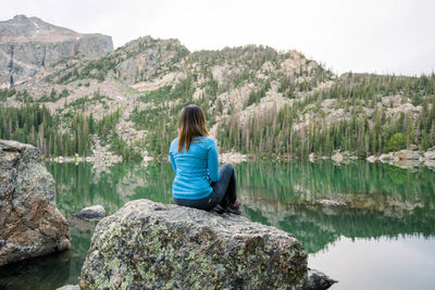 Rear view of woman sitting on rock by lake against mountains