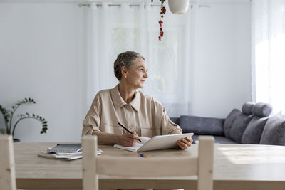 Thoughtful woman sitting at table at home with digital tablet and notebook