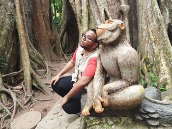 Young man sitting by statue against tree trunk in forest