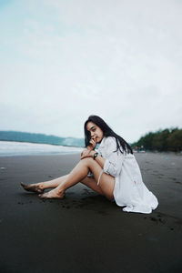 Portrait of young woman sitting on beach against sky