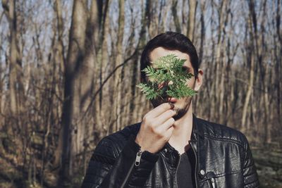 Close-up of man covering face with plants against bare trees in forest