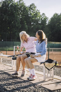 Happy senior female friends talking to each other while sitting on bench at tennis court