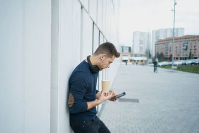 Side view of a modern man in casual clothes sitting near a white wall