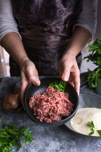 Unrecognizable female in apron demonstrating bowl with fresh minced meat and parsley to camera while cooking on gray table