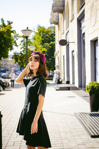 Beautiful young woman in black dress standing on sidewalk in city