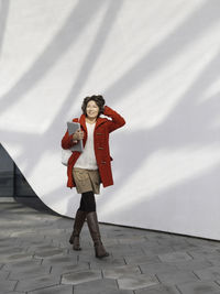 Curly woman in red duffle coat is walking by white wall. smiling student with laptop. 