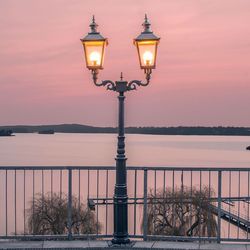 Illuminated street light by fence against lake during sunset