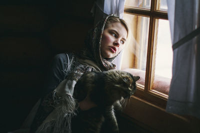 Thoughtful young woman with cat looking through window at home