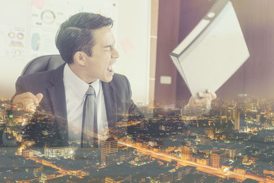 Double exposure of angry businessman holding file and illuminated cityscape