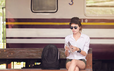 Young woman holding brochure while sitting on bench at railroad station 