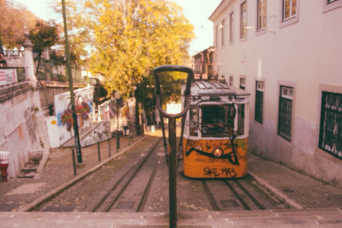 building exterior, architecture, built structure, transportation, tree, mode of transport, yellow, city, railroad track, street, land vehicle, car, outdoors, residential building, building, orange color, incidental people, residential structure, street light, day