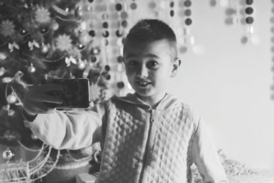 Portrait of smiling boy taking selfie while standing against christmas tree at home
