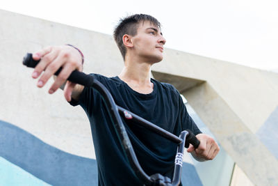 Low angle of young male in black t shirt touching bike handlebar and looking away while standing against concrete wall on weekend day in skate park