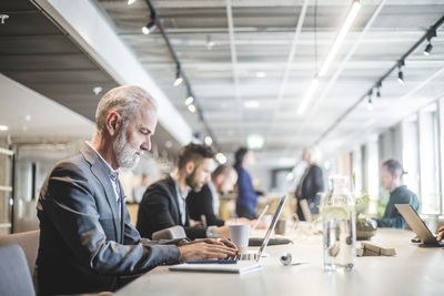 Confident senior businessman working over laptop while sitting with colleague at table in office