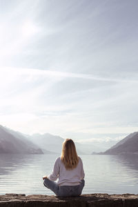 Rear view of woman sitting by lake against sky