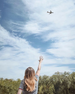 Rear view of woman gesturing towards airplane flying against sky