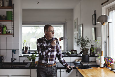 Senior man talking on smart phone through in-ear headphones while standing in kitchen at home