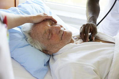 Cropped image of female nurse consoling senior man while male colleague examining with stethoscope in hospital ward