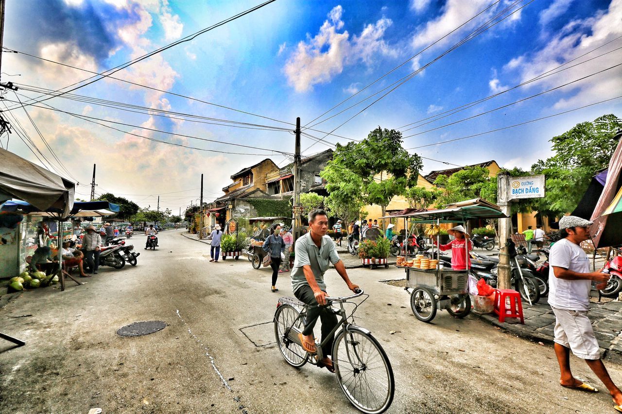 transportation, sky, mode of transport, men, bicycle, land vehicle, building exterior, street, lifestyles, tree, cloud - sky, person, architecture, built structure, leisure activity, cloud, large group of people, day, city