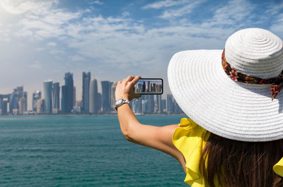 Rear view of woman photographing cityscape with smart phone by sea against sky