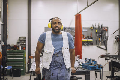 Portrait of smiling carpenter wearing ear protectors leaning on workbench in workshop