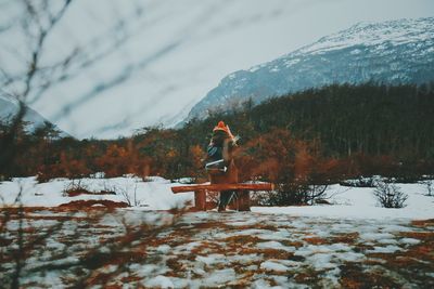 Rear view of woman wearing warm clothing while sitting on bench against mountains