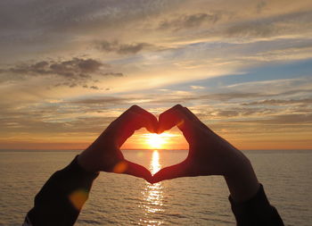Cropped hands making heart shape by sea against sky during sunset