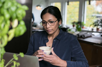 Businesswoman holding cup while looking at laptop in home office